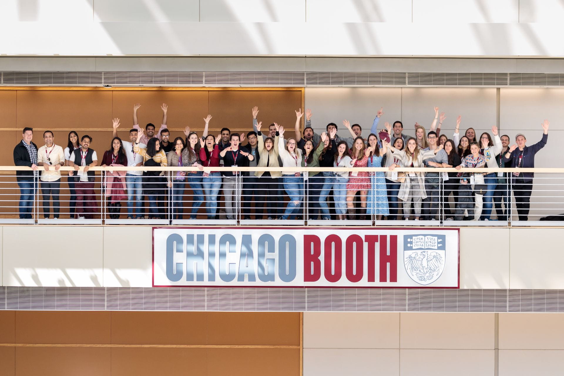 Group of people smiling and waving on an interior building balcony above a Chicago Booth sign