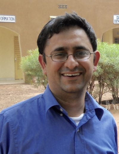 Kavi Bhalla PhD standing outside in a courtyard with trees. Kavi has short black hair and is wearing glasses and a blue collared shirt