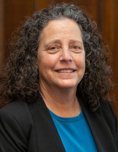 Leslie M. Kay, PhD is stilling in the SSRB tea room in front of a wood panel. She has long curly grey hair and is wearing a black blazer over a blue shirt