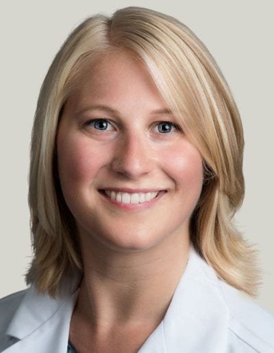 Angela M Lager, PhD sitting smiling in front of a light gray background. She is wearing a black blouse with a white medical coat over it. She has medium length blonde hair.