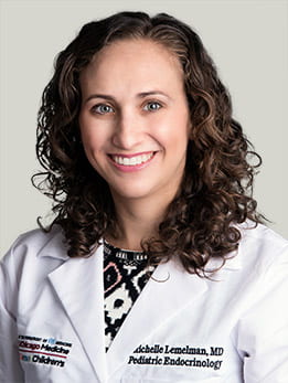 Michelle Lemelman, MD sitting smiling in front of a light gray background. She is wearing a black and white blouse with a white medical coat over it. She has long, curly brown hair.