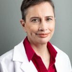 Iris Romero, MD, MS, sitting smiling in front of a medium gray background. She is wearing a red button-down shirt with a white medical coat over it. She has short brown hair.