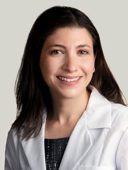 Mim Ari, MD sitting smiling in front of a light gray background. She is wearing a black blouse with a white medical coat over it. She has long brown hair.