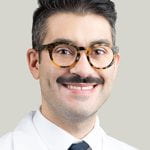 Daniel Z. Friedman, MD is smiling in front of a light gray background with a white collared shirt, blue tie, and white medical coat on. He has black short hair, a black mustache, and tortoise shell glasses on.