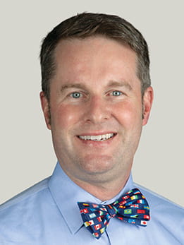 Peter J. Smith, MD, MA sitting smiling in front of a light gray background. He is wearing a light blue button-down shirt and a multicolor polka dot bowtie. He has brown short hair.