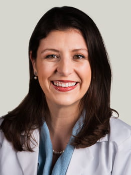 Sarah Sobotka, MD sitting smiling in front of a light gray background. She is wearing a blue blouse with a white medical coat over it. She has long brown hair.