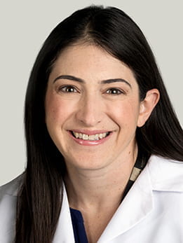 Melissa Tesher, MD sitting smiling in front of a light gray background. She is wearing a black and tan blouse with a white medical coat over it. She has long brown hair.