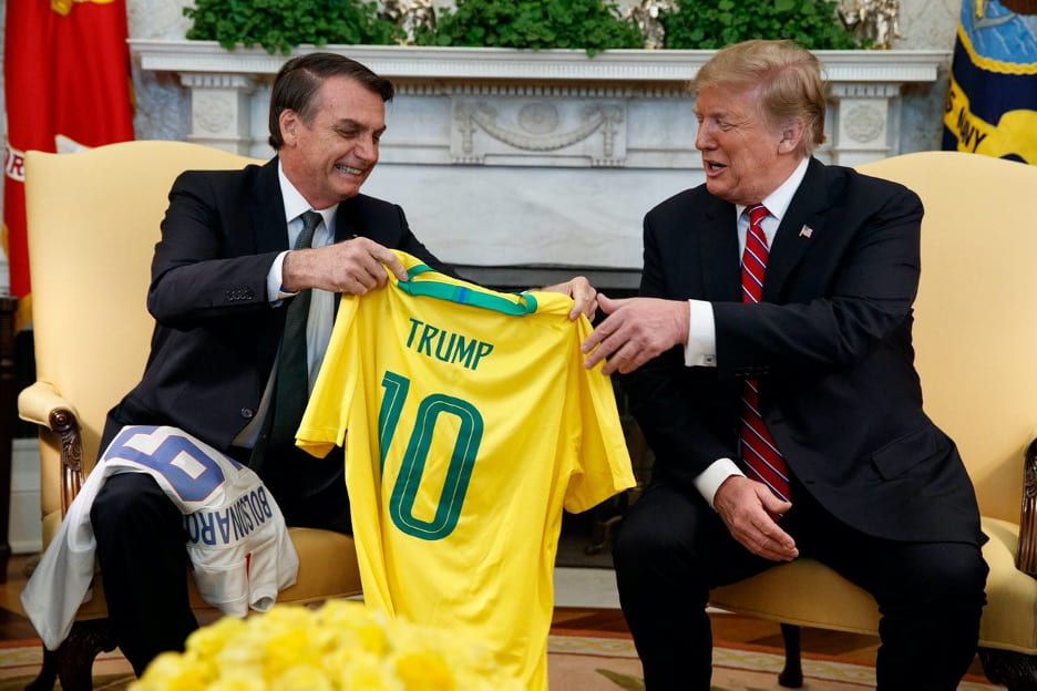 Jair Bolsonaro hands yellow soccer-style t-shirt to Donald Trump during a meeting.