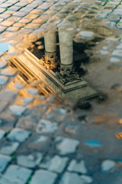 Photograph of pillars reflected in a pool of water on cobblestone.