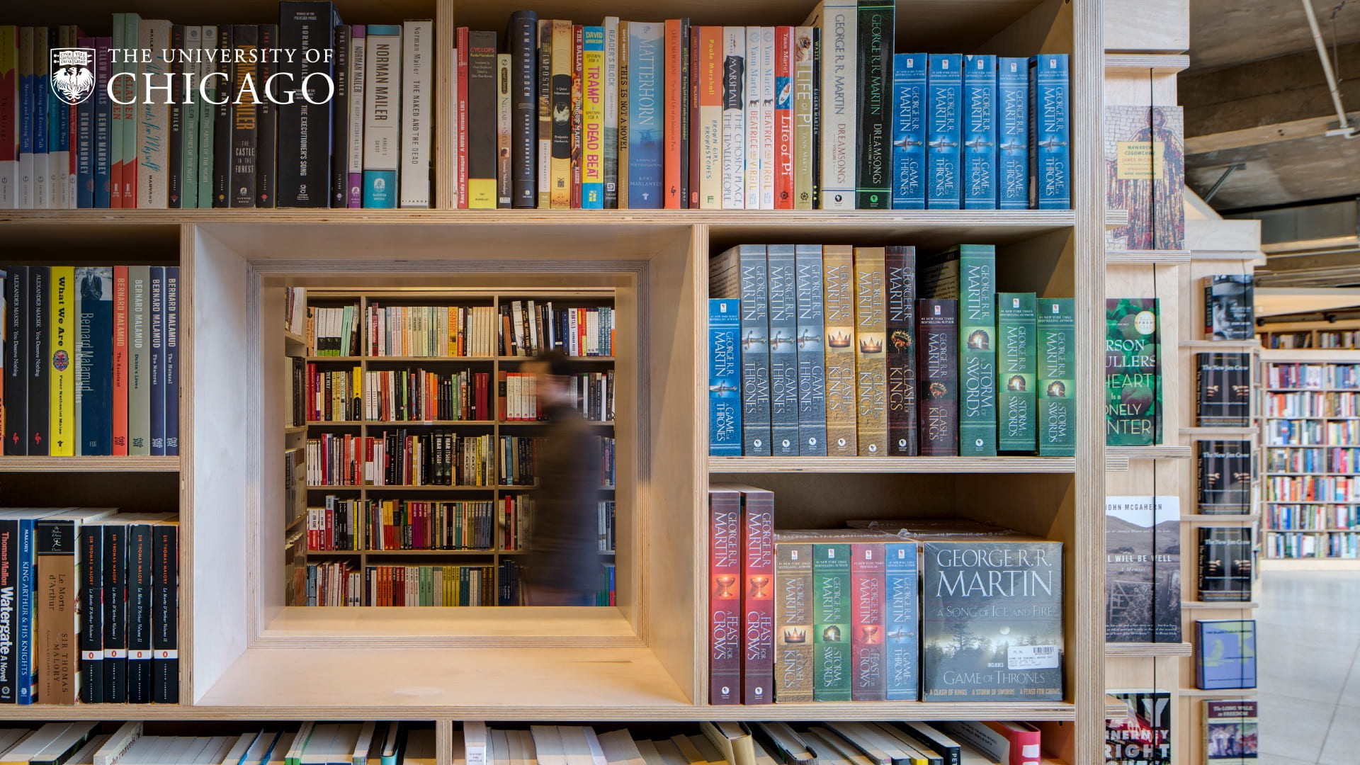 Looking through an opening in a bookshelf at a blurred person walking in the background